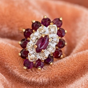 May include: A cluster ring featuring a central marquise-cut deep red gemstone surrounded by a halo of small round white stones, framed by an outer border of round deep red gemstones set in a silver-toned metal setting with gold prongs.