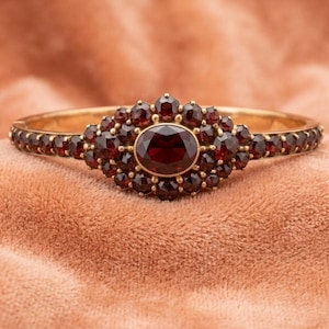 May include: A gold hinged bangle bracelet featuring a central oval-cut deep red garnet surrounded by a cluster of smaller round-cut garnets. The stones are set in a gold band that continues with a row of round garnets along the sides.