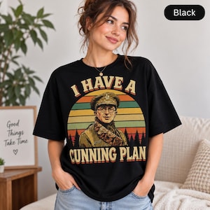 May include: A black t-shirt featuring a graphic print of a person in a military uniform and glasses. The design includes a sunset background with silhouettes of trees and the text "I HAVE A CUNNING PLAN" in a bold, retro-style font.
