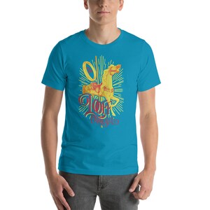 May include: A turquoise t-shirt featuring a carousel horse design in yellow and red. The horse is surrounded by a sunburst design, with the text "Loft Carousel" in a stylized font. The shirt is made of a soft material.
