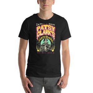 May include: Black t-shirt featuring a colorful graphic for "Patsy Clark's Haunted Mansion." The design includes the text "Can You Survive" and an illustration of a mansion with a person in front. The shirt is short-sleeved.