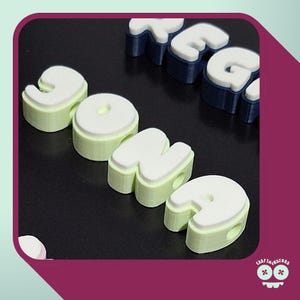 Könnte beinhalten: Ein Set aus 3D-gedruckten Buchstabenperlen, die den Namen JONA in Hellgrün mit weißen Oberseiten bilden und ein seitliches Loch zum Auffädeln aufweisen. Ein Teilwort, das mit REG beginnt, ist im Hintergrund in Dunkelblau mit weißen Oberseiten zu sehen.