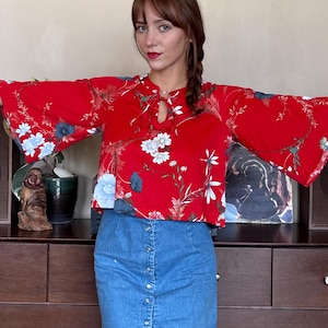 May include: A bright red blouse featuring a floral pattern in white, light blue, and dark green, with wide bell sleeves and a keyhole neckline with tie strings. Paired with a medium-wash blue denim skirt featuring a front row of silver metal buttons.