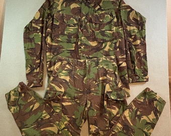 Può includere: Un completo composto da una camicia a maniche lunghe abbottonata e pantaloni coordinati con motivo mimetico nei toni del verde, marrone e beige. La camicia presenta due tasche sul petto con bottoni, mentre i pantaloni hanno due tasche cargo.