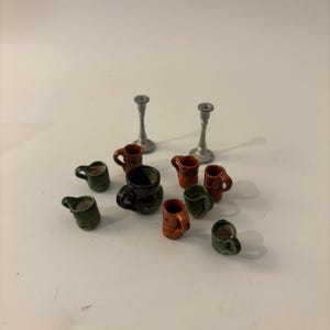 May include: A collection of miniature items including two silver-colored candlesticks, one large dark brown pitcher, and several small ceramic mugs in shades of green and brown, all arranged together on a plain surface for display purposes.