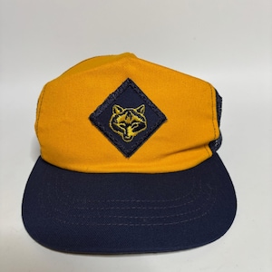 May include: A mustard yellow and navy blue trucker hat featuring a diamond-shaped patch on the front panel. The patch is navy blue with an embroidered yellow fox head graphic centered within it. The hat has a solid front and a mesh back.