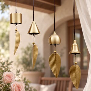 May include: Four hanging wind chimes with gold-toned metal components suspended by black cords. Each chime features a unique top shape including a cylinder, cone, dome, and bell, paired with a textured, leaf-shaped metal wind catcher below.