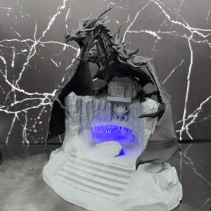 May include: A dark grey dragon figurine perched atop a light grey rocky base with a staircase. The base features a central alcove with a glowing blue light illuminating etched runic symbols. The dragon has large wings and an open mouth.