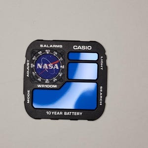以下が含まれることがあります： 円形のNASAロゴ、青いディスプレイパネル、および「5 ALARMS」、「WR100M」、「10 YEAR BATTERY」などの白いテキストが特徴の、黒いカシオ時計の文字盤交換パーツ。フレームには「ADJUST」、「LIGHT」、「MODE」、「SEARCH」の機能ラベルが含まれています。