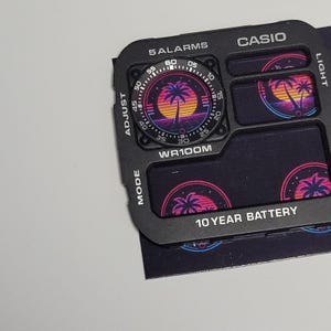 May include: A black Casio watch face replacement part featuring a retro sunset and palm tree graphic. The component includes white text labels for 5 ALARMS, LIGHT, ADJUST, WR100M, MODE, and 10 YEAR BATTERY, with a circular analog-style dial display.