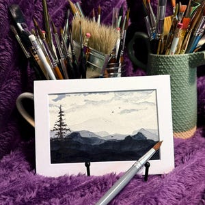 May include: A rectangular watercolor painting in a white mat frame depicts a landscape with a dark silhouette of a pine tree against layered blue and gray mountains under a cloudy sky. A gray paintbrush rests in front of the framed artwork.