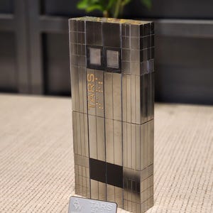 Peut inclure: Une figurine de robot rectangulaire et métallique composée de blocs segmentés avec une finition argentée réfléchissante. L'avant présente le texte TARS, deux panneaux carrés en forme d'yeux et une section rectangulaire noire. Une plaque indique TARS.