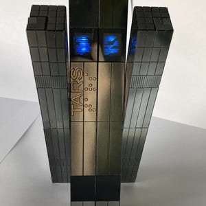 May include: A metallic, block-like robot figure composed of dark gray, segmented rectangular panels. The front panel features the text TARS in vertical lettering, accompanied by a grid of small circular indentations and two glowing blue screens.