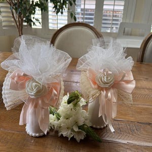 May include: Two decorative white and peach-colored favors with tulle, ribbons, and a rose embellishment. The favors are displayed on a wooden table, alongside a small bouquet of white flowers. The favors are decorated with pearls.