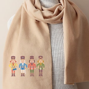 Op de afbeelding: Een beige sjaal met een ontwerp van vier notenkrakersoldaten in kleurrijke uniformen. De sjaal is gevouwen en gedrapeerd over een witte trui.
