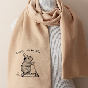 Könnte beinhalten: Beigefarbener Schal mit einer Illustration eines Bibers auf einem Roller, der einen Zaubererhut trägt. Der Text "Off to cause a kerfuffle" ist aufgedruckt. Der Schal ist über einer Schaufensterpuppe drapiert.