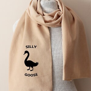 Op de afbeelding: Een beige sjaal met een zwart silhouet van een gans en de tekst "Silly Goose" erop gedrukt.