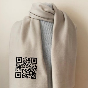 May include: A light beige scarf draped over a mannequin. The scarf has a QR code printed in black on the lower left side. The scarf is made of a soft material and is wrapped around the mannequin's neck.