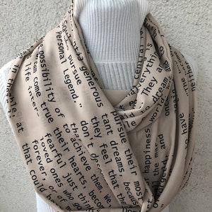 May include: Beige infinity scarf with black text. The scarf is draped over a white ribbed turtleneck. The text includes phrases like "pursue their dreams" and "Personal Legend."