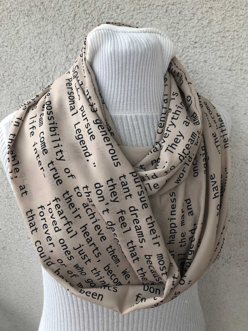 May include: Beige infinity scarf with black text. The scarf is draped over a white ribbed turtleneck. The text includes phrases like "pursue their dreams" and "Personal Legend."