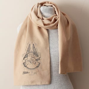May include: A beige scarf displaying a black blueprint of the Millennium Falcon, with the text "Corellian Engineering Corporation". The scarf is draped over a grey sweater.