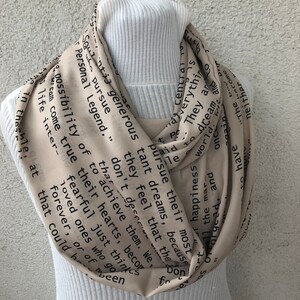 May include: A beige infinity scarf with black text. The scarf is draped around a white turtleneck. The text includes phrases like "pursue their most tant dreams" and "Personal Legend."