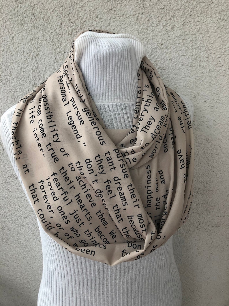 May include: A beige infinity scarf with black text. The scarf is draped around a white turtleneck. The text includes phrases like "pursue their most tant dreams" and "Personal Legend."