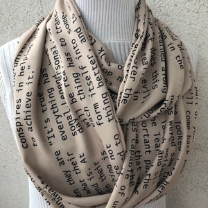 May include: Beige infinity scarf with black text. The scarf is wrapped around a white turtleneck. The text appears to be a quote or passage from a book. The scarf is made of a lightweight material.