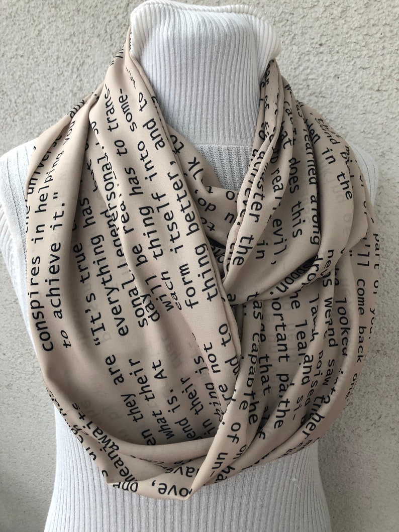 May include: Beige infinity scarf with black text. The scarf is wrapped around a white turtleneck. The text appears to be a quote or passage from a book. The scarf is made of a lightweight material.