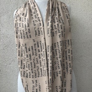 May include: A beige infinity scarf with black text. The scarf is draped around a white turtleneck. The text appears to be a poem or a series of quotes. The scarf is made of a soft, flowing material.