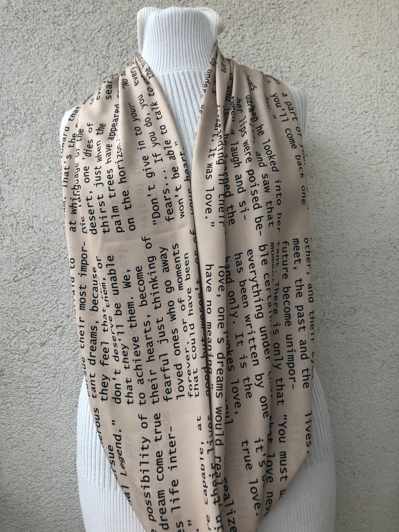 May include: A beige infinity scarf with black text. The scarf is draped around a white turtleneck. The text appears to be a poem or a series of quotes. The scarf is made of a soft, flowing material.