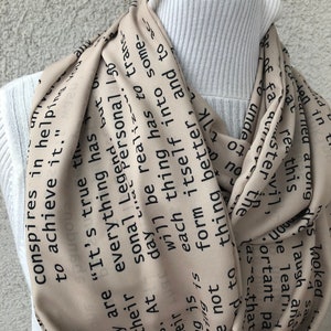 May include: A beige infinity scarf with black text. The scarf is draped around a white turtleneck. The text includes phrases like "It's true" and "conspires in helping to achieve it."