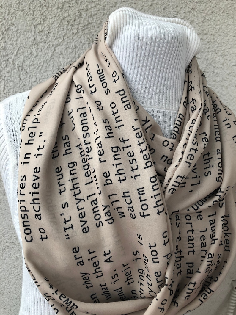 May include: A beige infinity scarf with black text. The scarf is draped around a white turtleneck. The text includes phrases like "It's true" and "conspires in helping to achieve it."