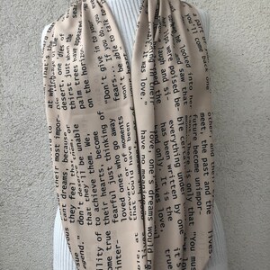 May include: A beige scarf with black text, draped over a white turtleneck. The text appears to be excerpts from a poem or story. The scarf is made of a lightweight material and has a flowing design.