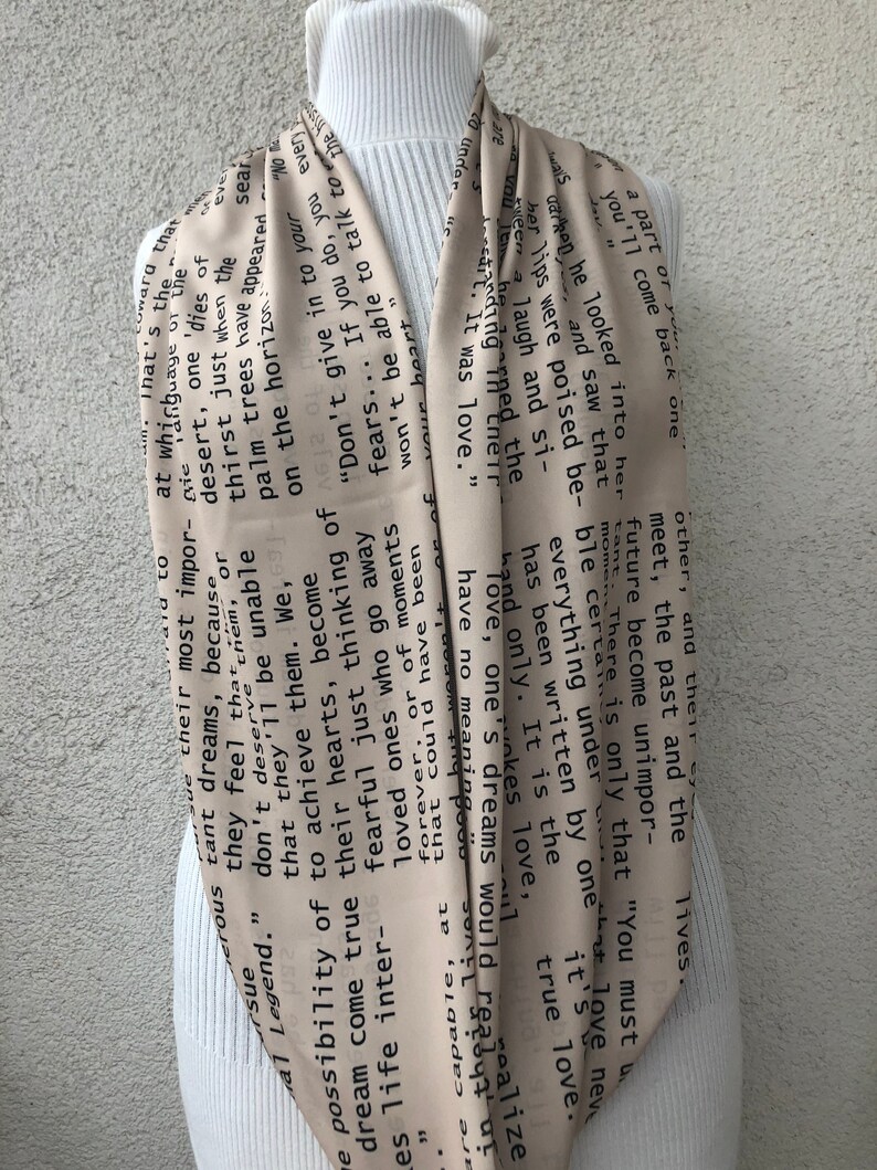 May include: A beige scarf with black text, draped over a white turtleneck. The text appears to be excerpts from a poem or story. The scarf is made of a lightweight material and has a flowing design.