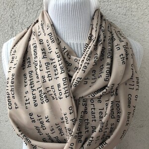 May include: A beige infinity scarf with black text. The scarf is draped around a white turtleneck. The text includes phrases like "It's true that everything has to come," and "Conspires to achieve it."