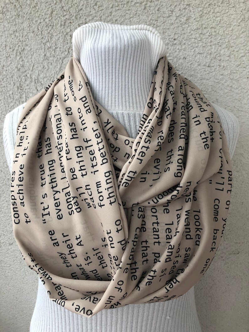 May include: A beige infinity scarf with black text. The scarf is draped around a white turtleneck. The text includes phrases like "It's true that everything has to come," and "Conspires to achieve it."