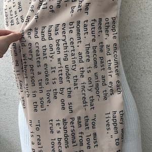 May include: A beige scarf with black text, draped over a mannequin. The text appears to be excerpts from a book or poem. The scarf is a neutral color, making it versatile for various outfits.