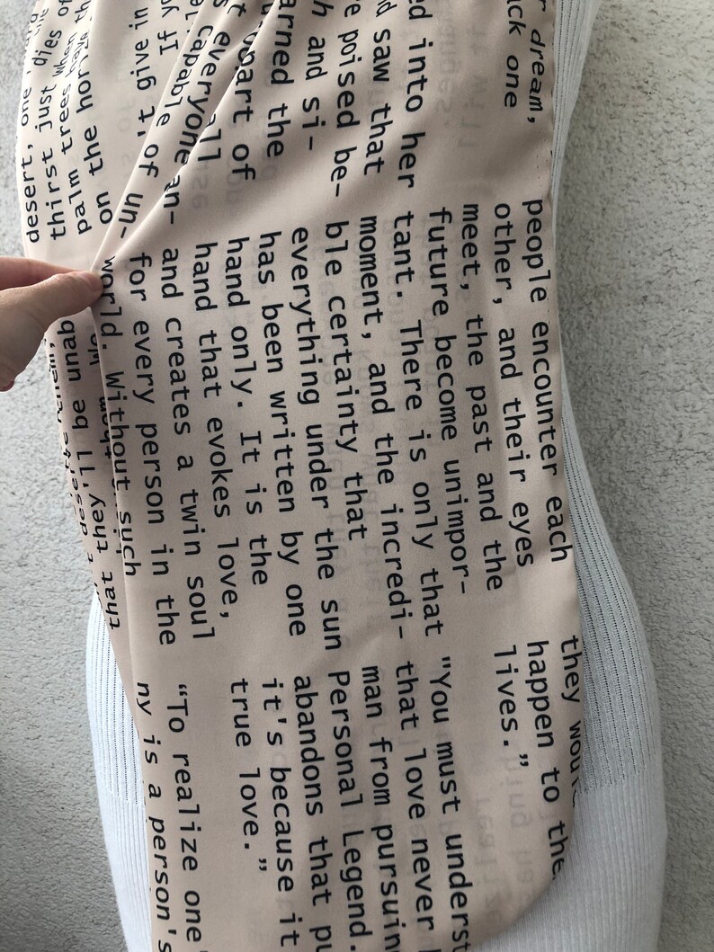 May include: A beige scarf with black text, draped over a mannequin. The text appears to be excerpts from a book or poem. The scarf is a neutral color, making it versatile for various outfits.