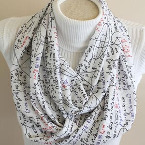 May include: A white infinity scarf with black and blue mathematical equations and formulas printed on it. The scarf is worn around a white turtleneck.
