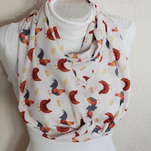 May include: White infinity scarf with a repeating pattern of colorful roosters. The roosters are in shades of red, orange, yellow, and black. The scarf is draped around a white turtleneck.