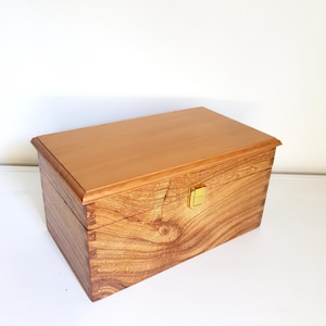 May include: A rectangular wooden box with a smooth, light-toned lid and a darker, grained wood body featuring visible wood knots. The box has a hinged lid with a small, centred gold-coloured metal latch on the front for closure.
