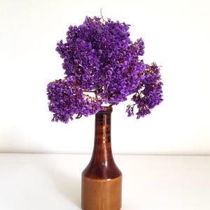 May include: A tall, slender brown ceramic bottle with a glossy finish serves as a vase for a large, dense cluster of dried purple statice flowers. The flowers feature numerous small, vibrant violet petals arranged in a rounded, bushy shape.