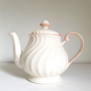 May include: A cream-coloured ceramic teapot featuring a swirling, ribbed texture on the body and lid. The teapot is accented with delicate pink trim along the lid edge, the handle, the spout, and the small knob on top of the lid.
