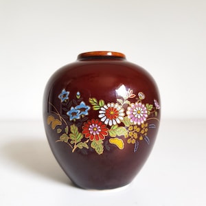 Può includere: Un vaso in ceramica color marrone scuro dalla forma bulbosa con un motivo floreale dipinto a mano. Il design presenta grappoli di crisantemi rossi, bianchi e rosa insieme a piccoli fiori blu a campana, foglie verdi e dettagli dorati.