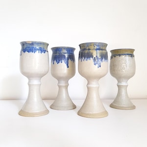 May include: A set of four ceramic goblets with a speckled off-white base and a flared pedestal foot. Each goblet features a distinct blue glaze dripping from the rim down the upper bowl, creating a textured contrast against the matte finish.