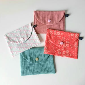 May include: Four fabric pouches with different patterns and colors. The pouches have a flap with a button closure. The patterns include a pink and white geometric pattern, a red floral pattern, a solid teal color, and a solid pink color.