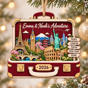 May include: A wooden suitcase-shaped ornament featuring the text Emma & Noah's Adventure and 2026. The design displays landmarks including the Eiffel Tower, Big Ben, Mount Fuji, the Statue of Liberty, and the Colosseum, with a signpost listing city names.