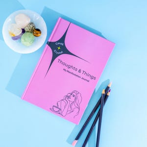 May include: A bright pink journal titled Thoughts & Things My Manifestation Journal with a black star graphic and line art of a person. Beside it sits a white dish containing several polished multi-colored crystals and three dark colored pencils.