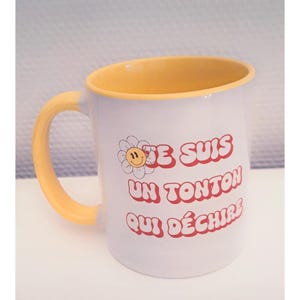 Peut inclure: Un mug en céramique blanc avec un intérieur et une anse jaunes. L'extérieur présente un motif de fleur marguerite souriante et le texte rouge "JE SUIS UN TONTON QUI DÉCHIRE" écrit dans une police grasse, arrondie et rétro sur la face.